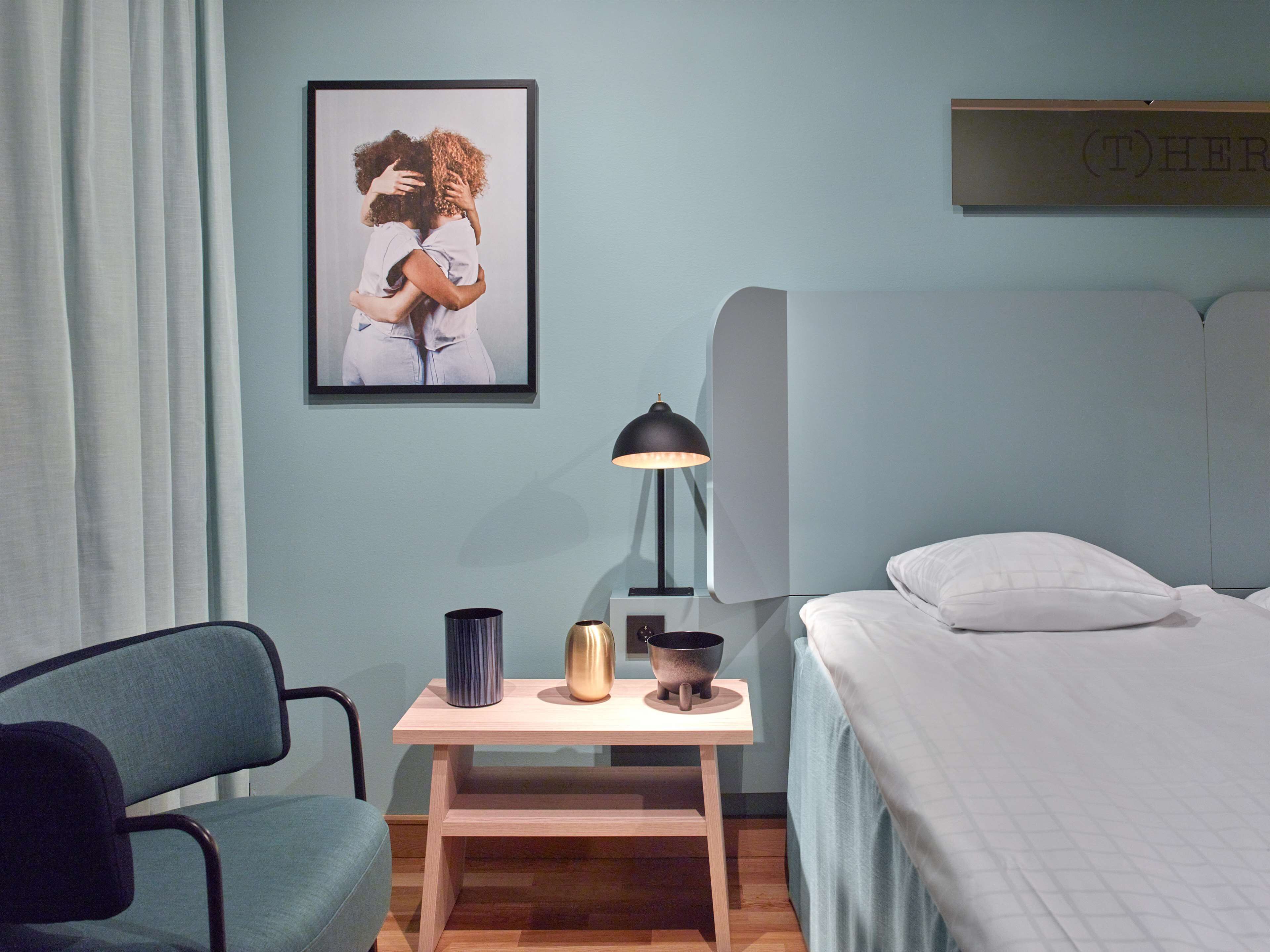 Un dormitorio con una cama, una mesita de noche con lámpara, una silla e imagen enmarcadas en la pared.