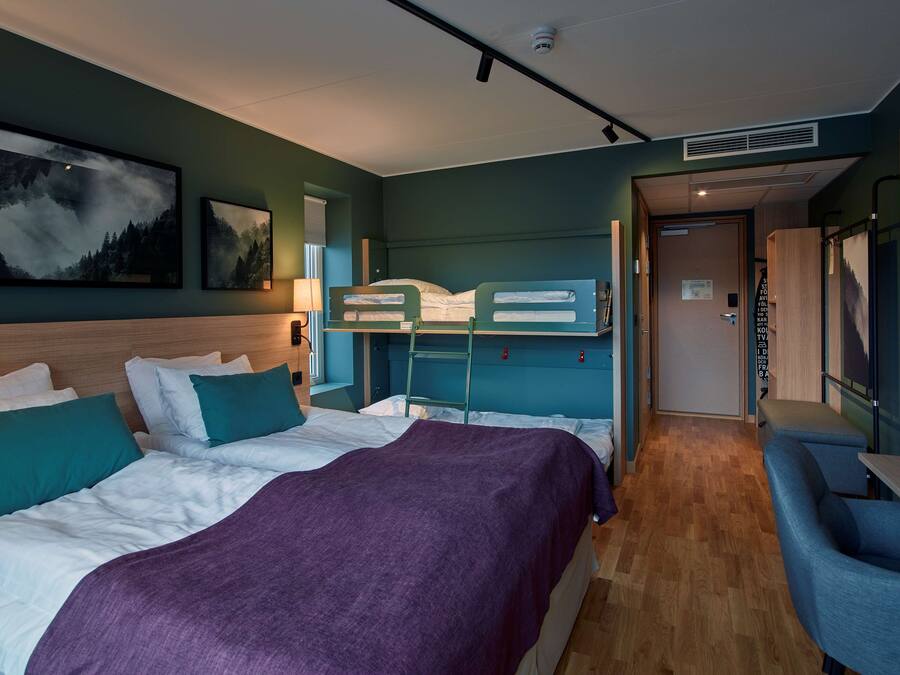 A hotel room with a bunk bed, a single bed, a purple blanket, a desk, and a chair.