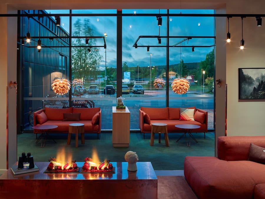 Un interior moderno con sofás rojos, una mesa de centro con chimenea integrada y ventanas amplias que dan a un estacionamiento y árboles.