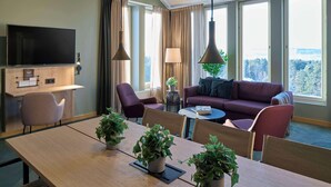 A modern hotel room with a wooden desk, a flat-screen TV, a sofa, and a dining table with chairs. There are potted plants on the table and a view of the outdoors through large windows.