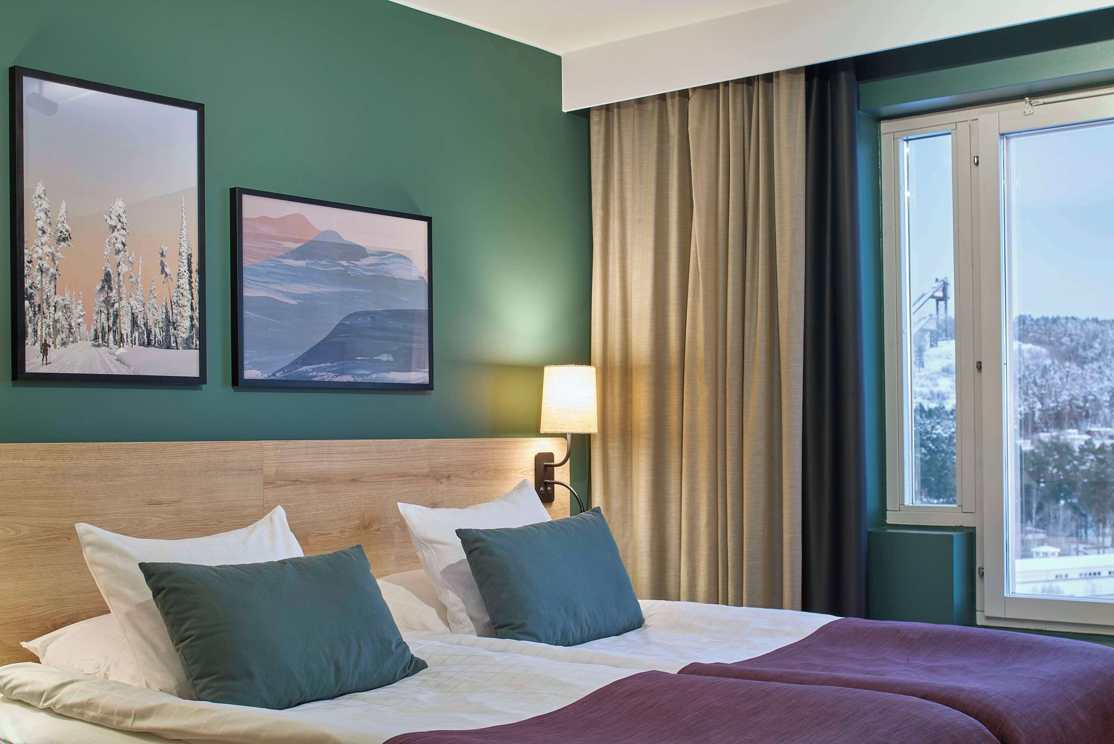 A hotel room with a large bed, two pillows, a headboard, a window with curtains, and two framed pictures on the wall.