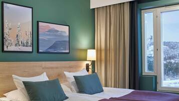 A hotel room with a large bed, two pillows, a headboard, a window with curtains, and two framed pictures on the wall.