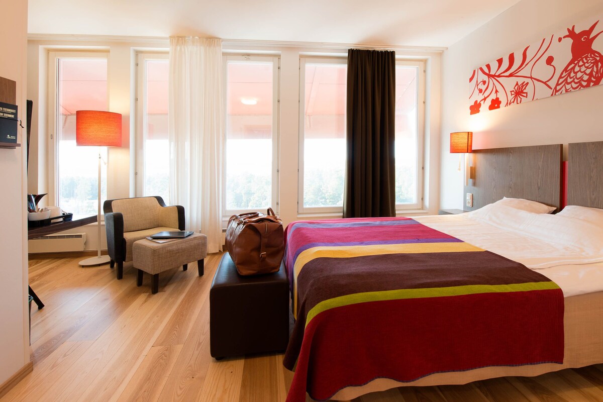 A hotel room with a bed, a sofa, a desk with a lamp, a suitcase, and a window with curtains.