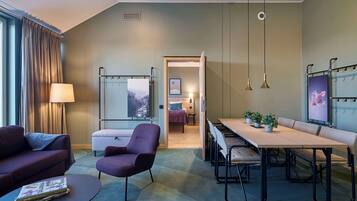 A modern hotel room with a large bed, a desk, a dining table, and a sofa. There is a window with curtains, a lamp, and a picture on the wall.