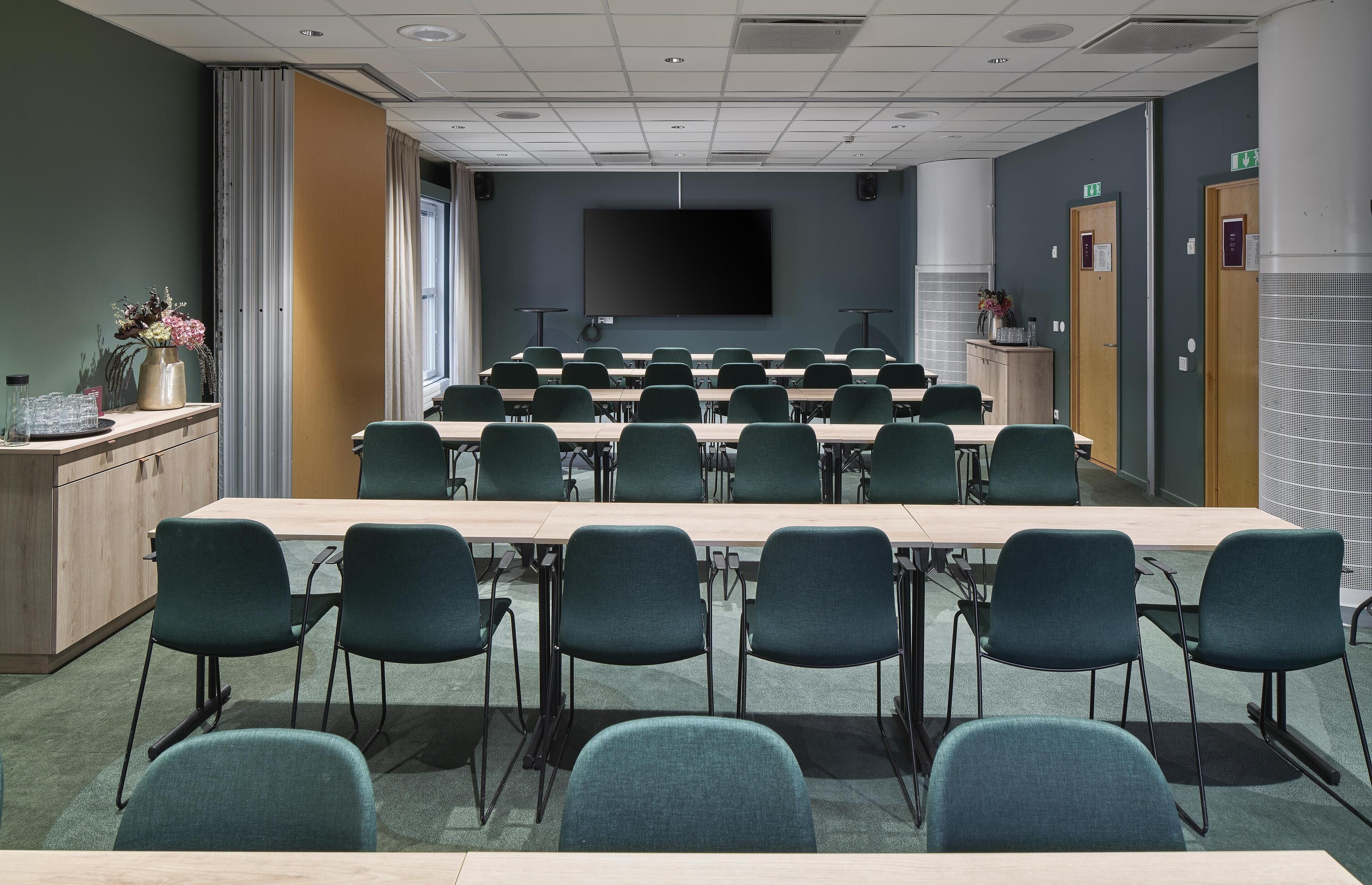 A conference room with a long table, chairs, a screen, speakers, and a small cabinet with items.