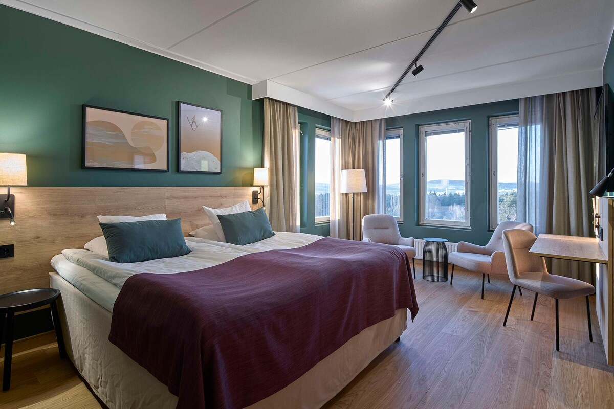 A hotel room with a large bed, two bedside tables, a desk, and a chair. There are two windows with curtains, a flat-screen TV, and artwork on the walls.