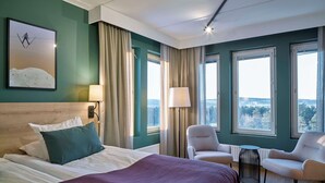 A hotel room with a bed, two chairs, a small table, a lamp, and a large window offering a view.