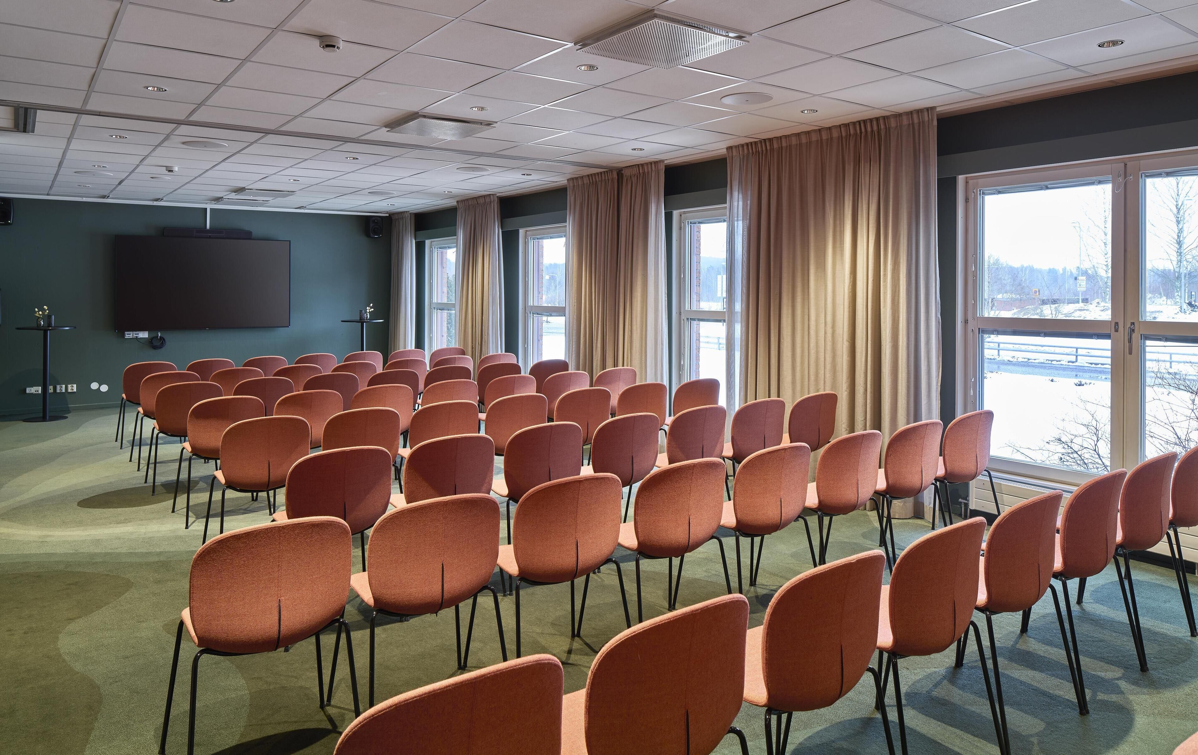 Uma sala de conferências com fileiras de cadeiras, uma tela de projetor e grandes janelas com cortinas.