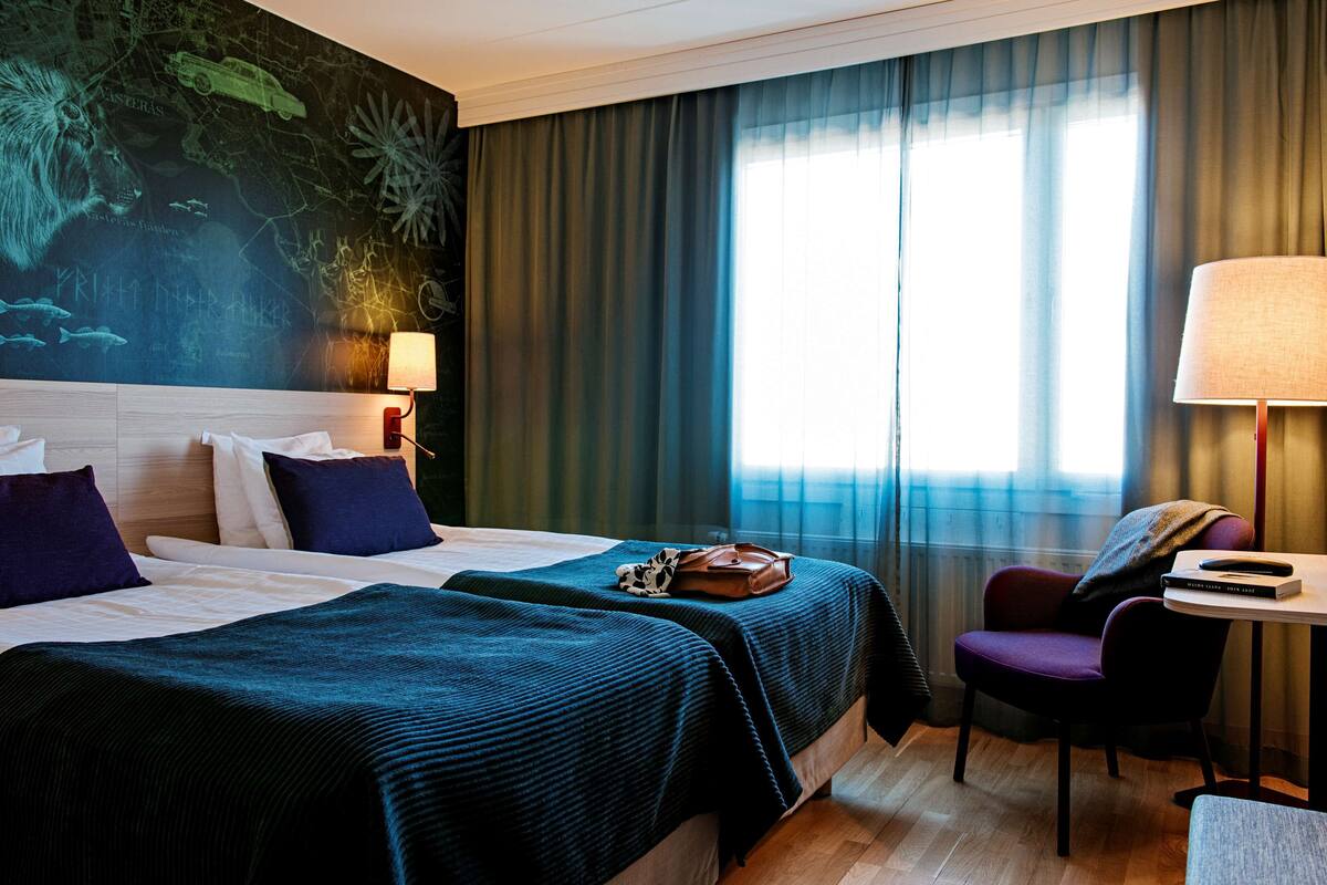 A hotel room with two beds, a large window with curtains, a chair, a table, and a wall with a map design.