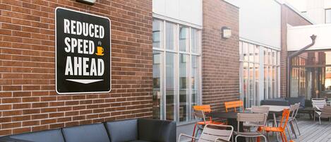 An outdoor seating area with a black leather sofa, orange chairs, and a red table. A sign on the wall reads "REDUCED SPEED AHEAD."