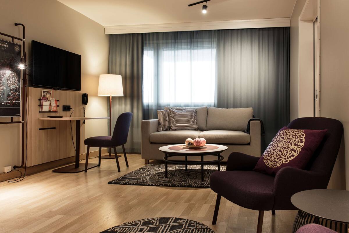 A modern living room with a sofa, armchairs, a coffee table, a TV, and a desk with a lamp.