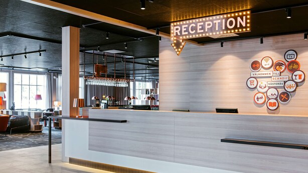 A reception area with a sign indicating 'RECEPTION' and a wall with various icons representing amenities such as Wi-Fi, TV, and a bicycle. - Scandic Västerås (Vasteras)