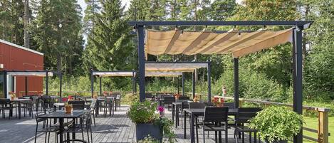 An outdoor patio with wooden tables and chairs, covered by pergolas, surrounded by greenery and trees.