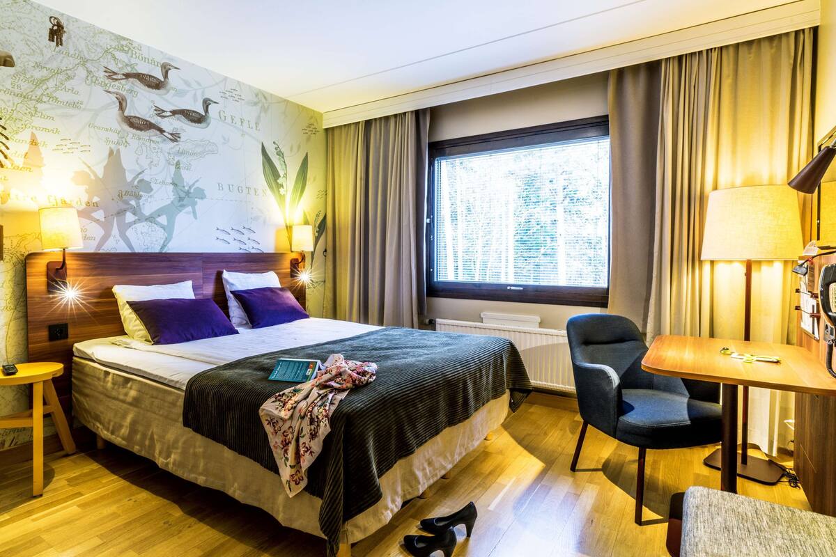 A hotel room with a large bed, a desk with a lamp, a chair, and a window with curtains.