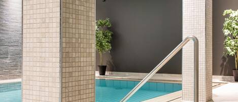 An indoor swimming pool with tiled walls, a metal handrail, and potted plants.