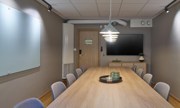 A conference room with a long table, chairs, a whiteboard, a projector screen, and a ceiling light fixture.