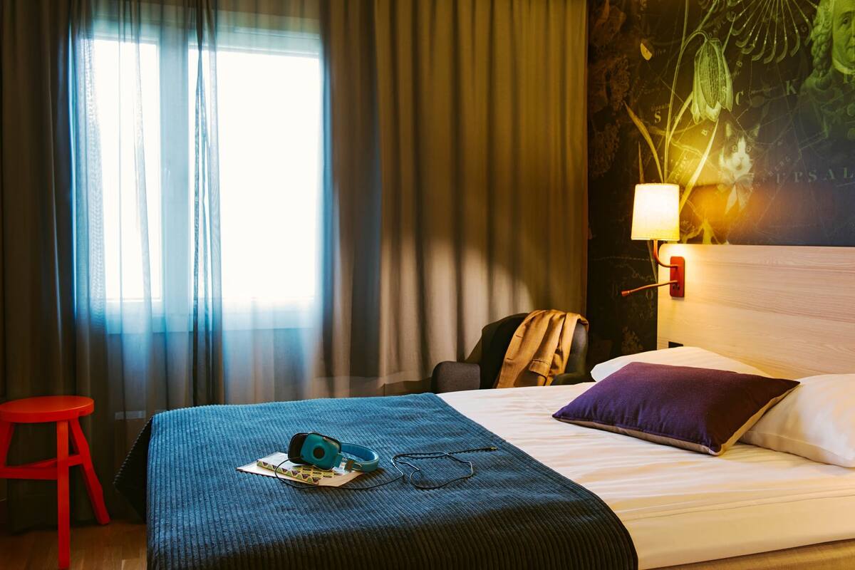 A hotel room with a bed, a red stool, a lamp, and a wall with a botanical mural.