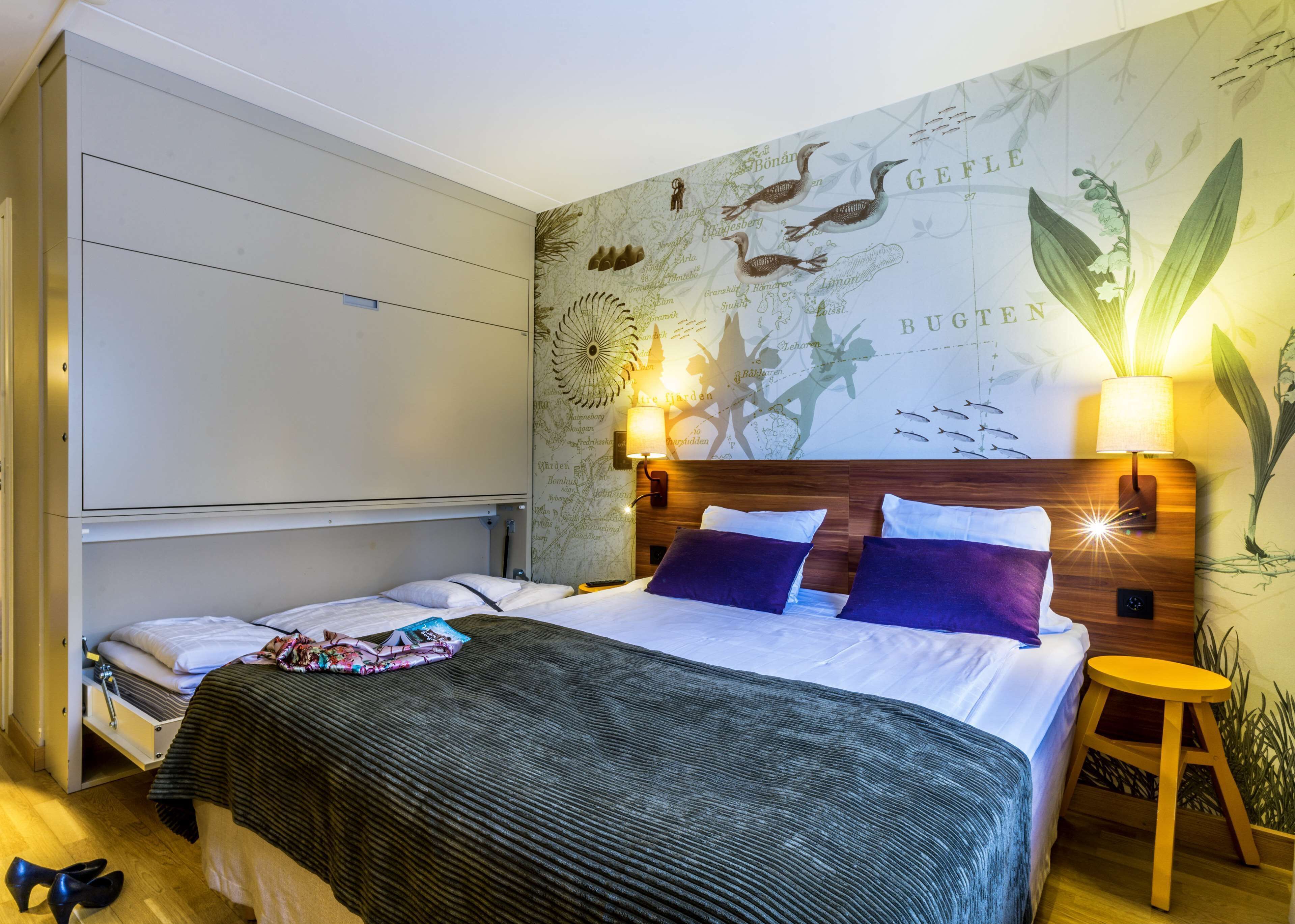 Habitación de hotel con cama, mesita de noche, lámpara y pared decorada con ilustraciones de un mapa y plantas.