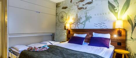 A hotel room with a bed, a nightstand, a lamp, and a wall with a map and plant illustrations.