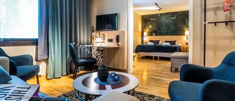 A modern hotel room with a seating area, a desk, a bed, and a TV.