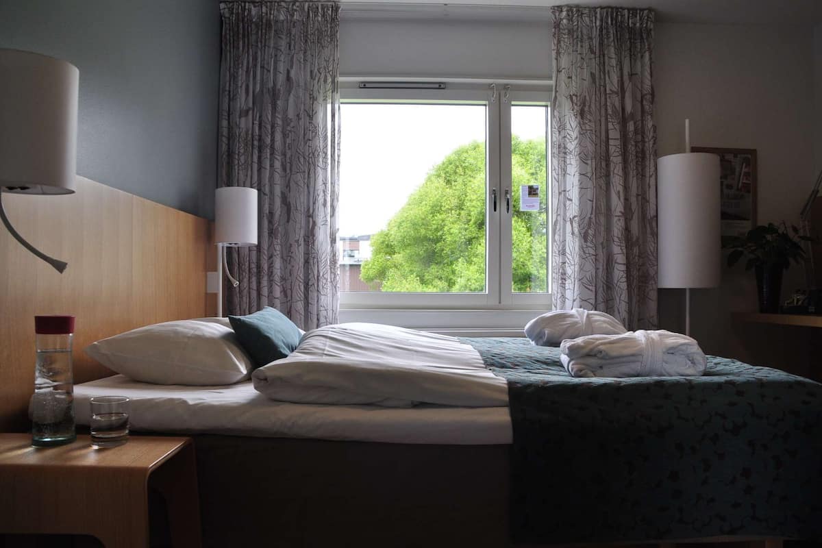 A hotel room with a bed, two bedside lamps, a window with curtains, a bedside table with a bottle and glass, and a wall-mounted picture frame.