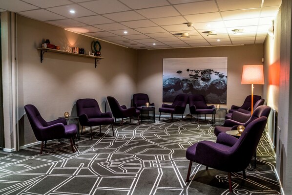 Uma sala de conferências com carpete estampado em padrão geométrico, cadeiras roxas, uma mesa redonda e uma prateleira fixada na parede com objetos decorativos.