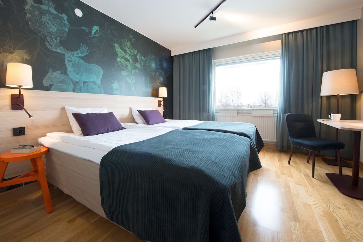 A hotel room with two beds, a desk, a chair, and a large window.
