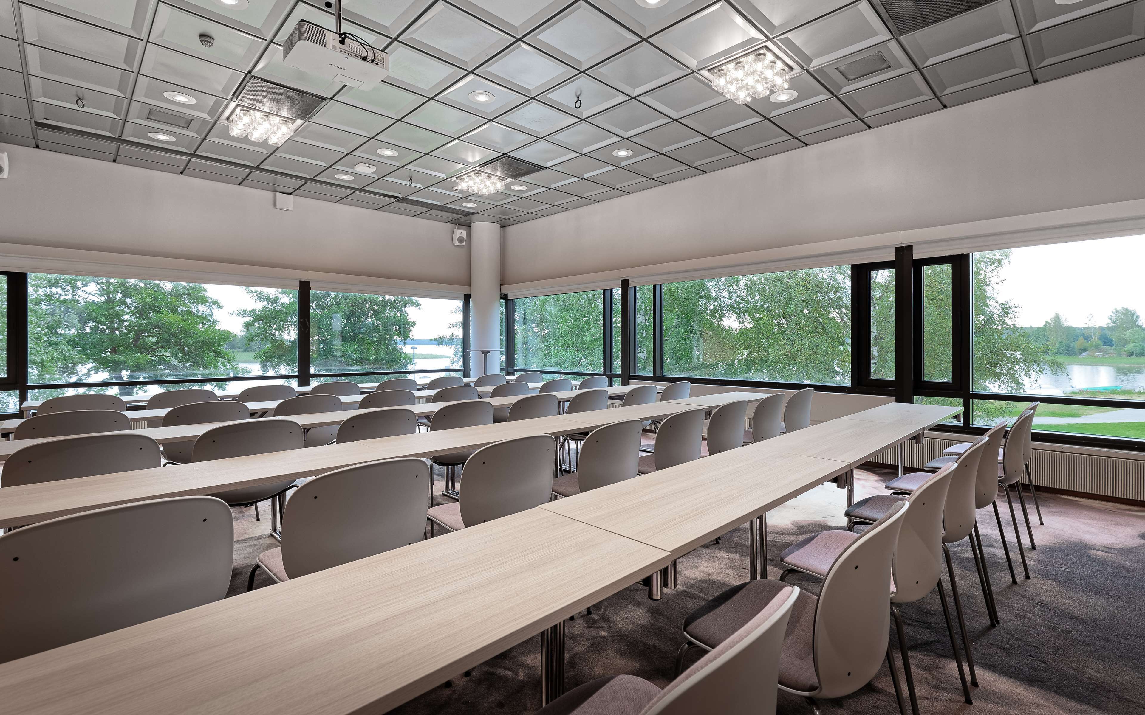 A conference room with long tables and chairs, large windows with a view of trees and a lake, and a ceiling with recessed lighting and a projector.