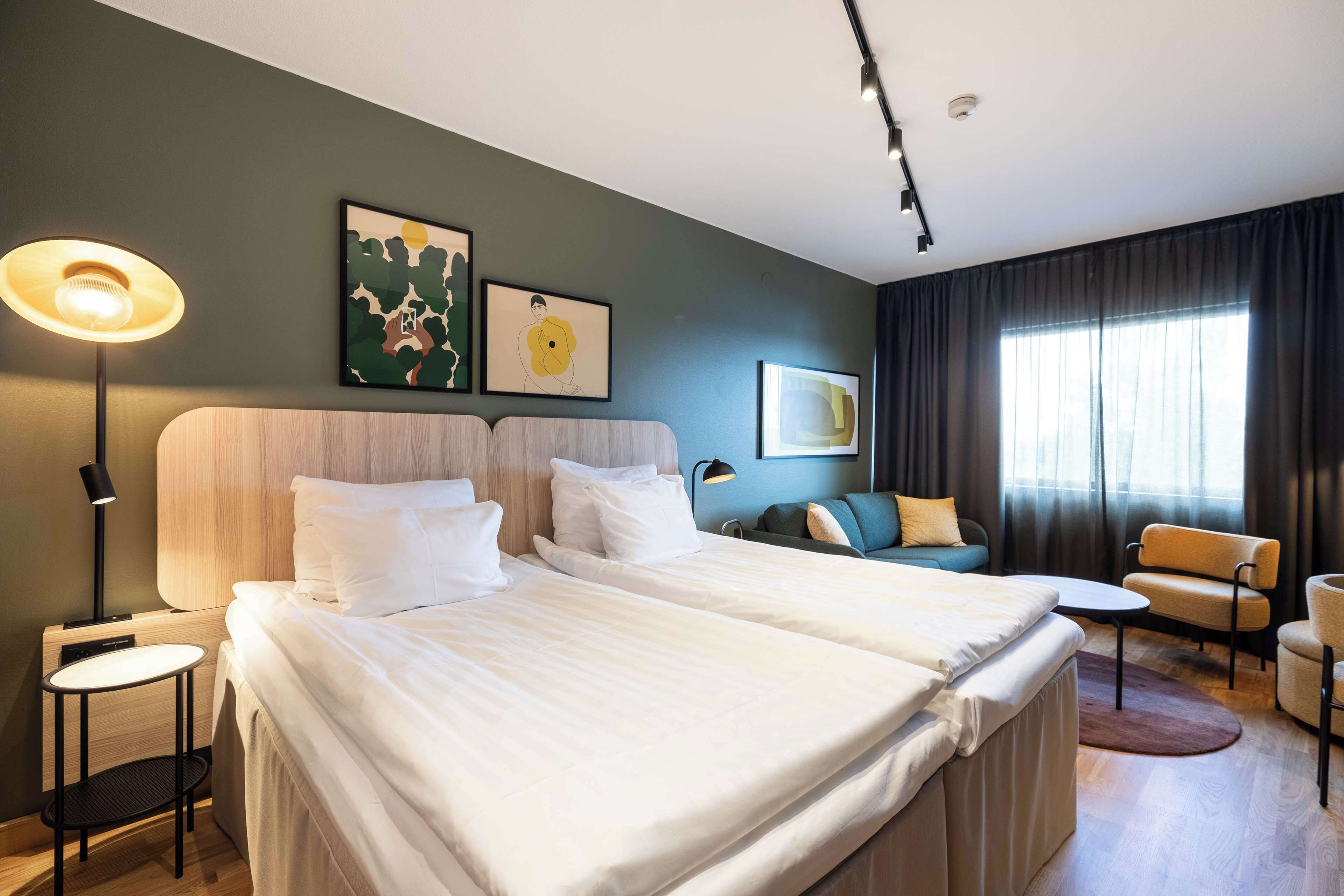 A hotel room with two beds, a desk, a chair, and a sofa. There are two wall-mounted lamps and artwork on the wall.