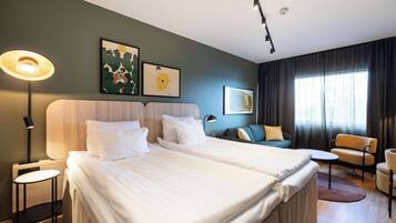 A hotel room with two beds, a desk, a chair, and a sofa. There are two wall-mounted lamps and artwork on the wall.