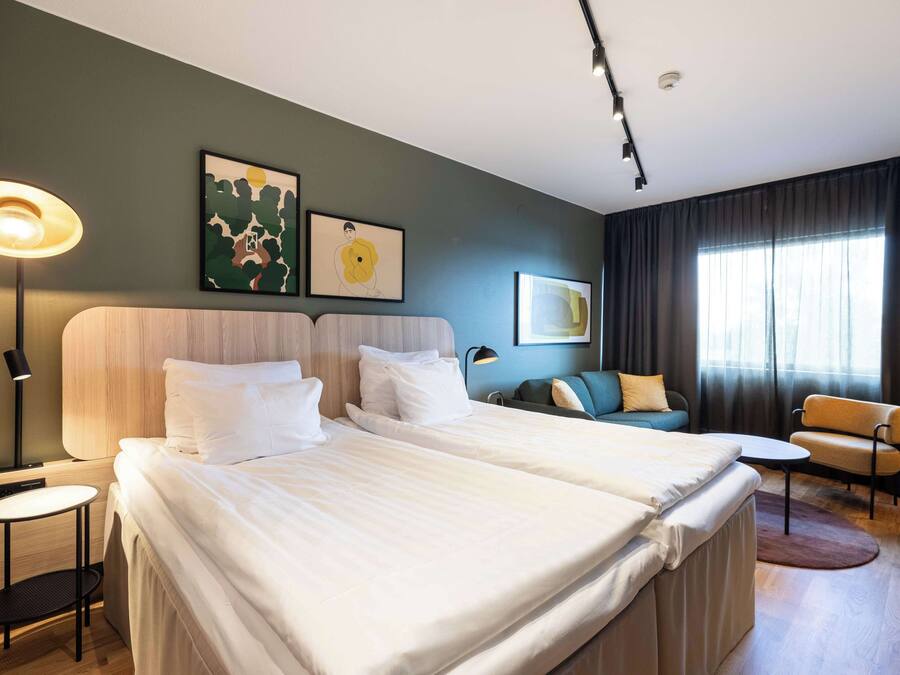 A hotel room with two beds, a desk, a chair, and a sofa. There are two wall-mounted lamps and artwork on the wall.
