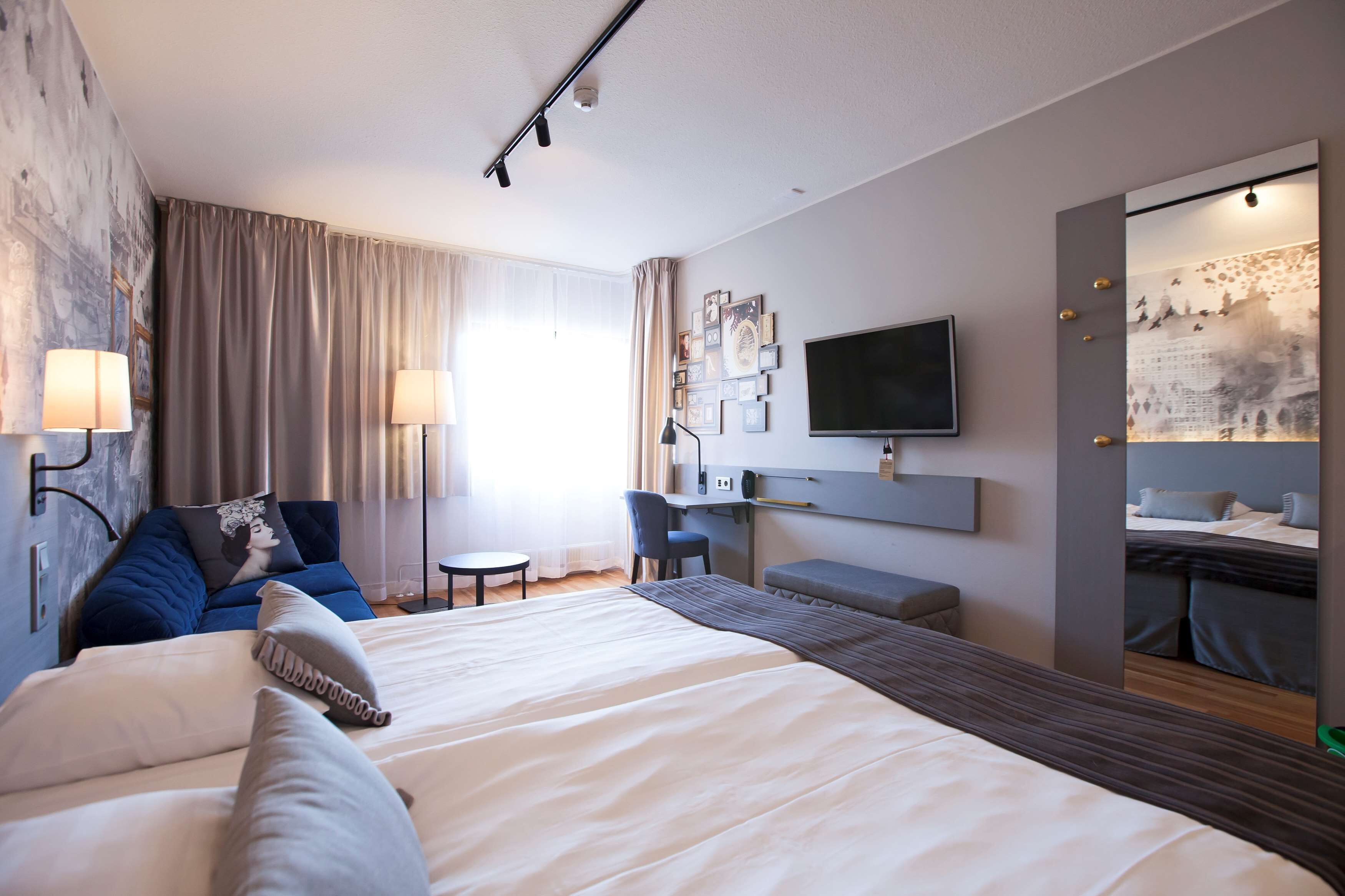A modern hotel room with a large bed, a desk, a chair, a TV, and a wall-mounted mirror.
