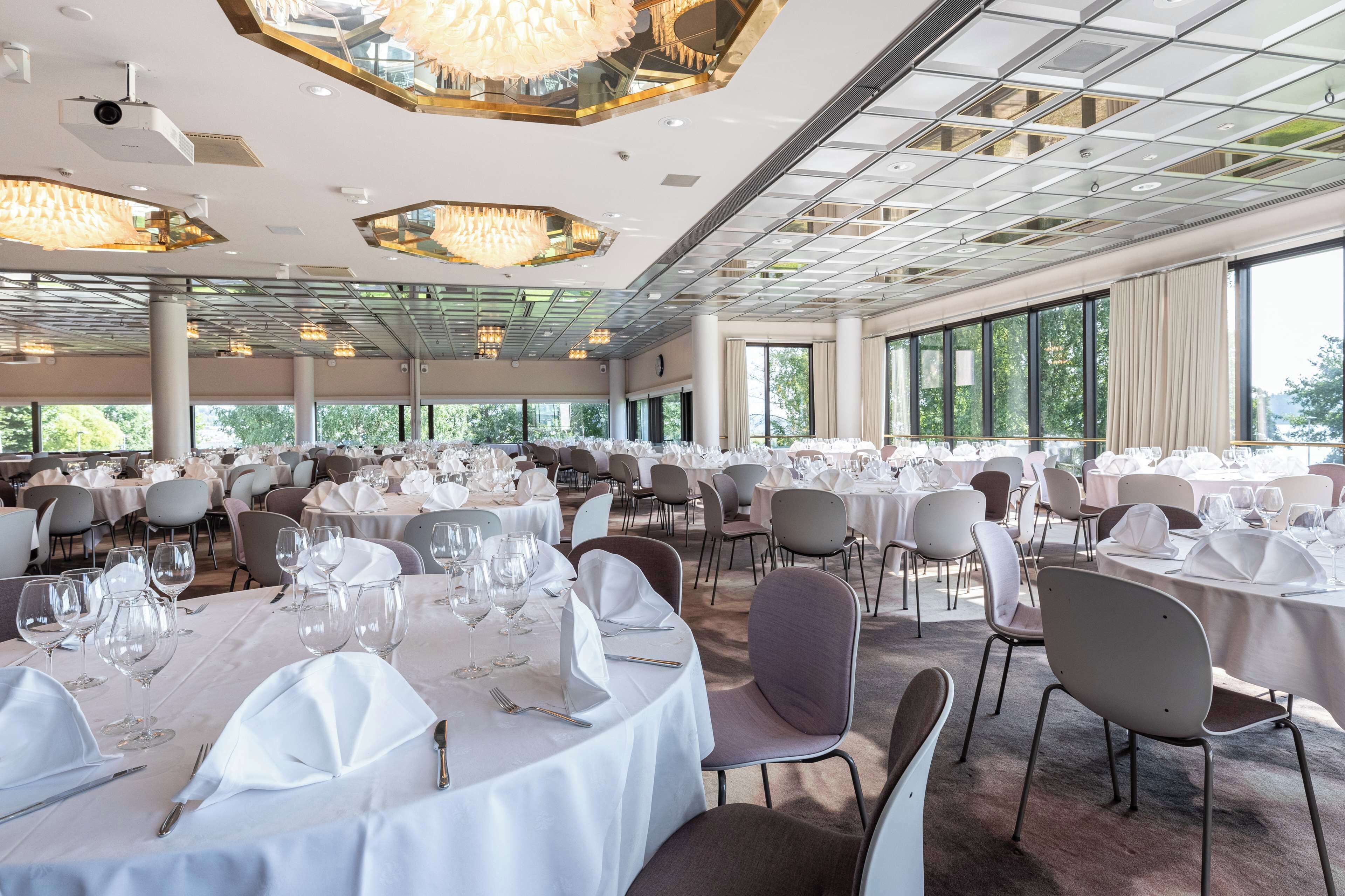 Une salle de banquet spacieuse, avec des tables rondes dressées pour le repas, de grandes fenêtres et des lustres modernes.