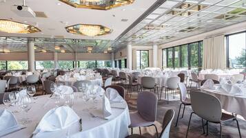 Une salle de banquet spacieuse, avec des tables rondes dressées pour le repas, de grandes fenêtres et des lustres modernes.