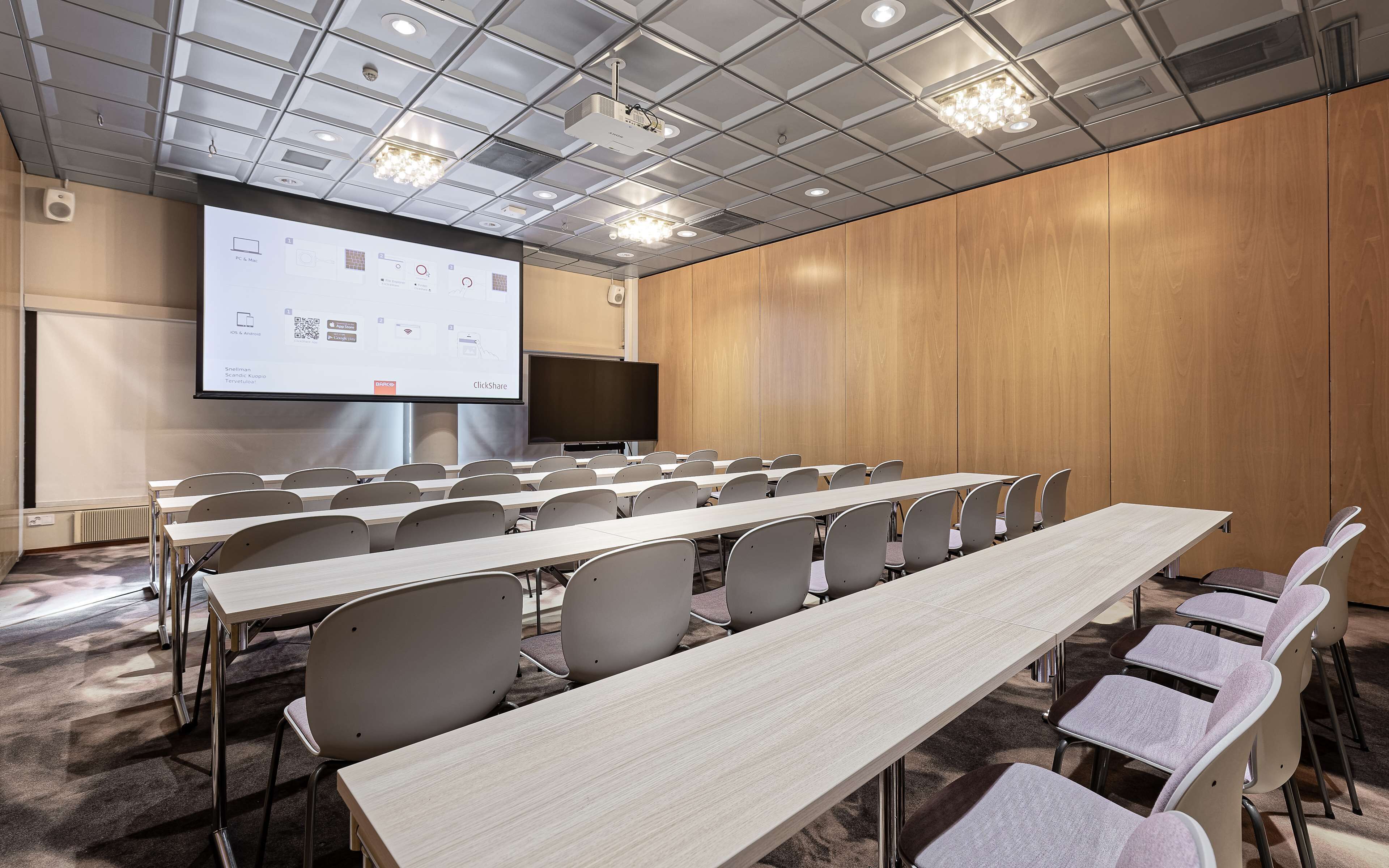 Un salón de conferencias con mesas largas y sillas, una pantalla de proyector y una pantalla grande para presentaciones.