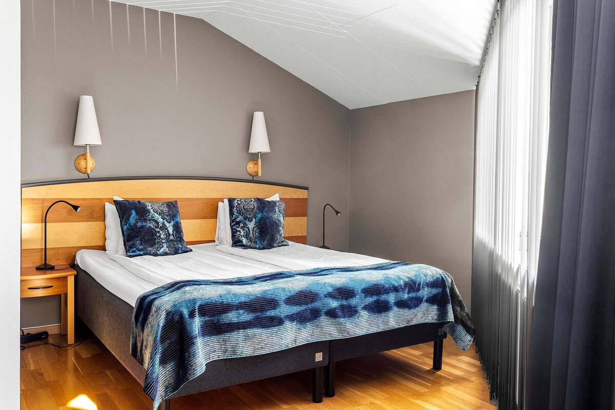 Une chambre d’hôtel moderne avec un grand lit, deux lampes de chevet, une tête de lit avec un revêtement en bois et une vue sur la ville par la fenêtre.