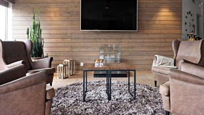 Ein modernes Wohnzimmer mit großem Flachbildfernseher an einer Holzwand, Ledersofas, einem Glastisch und einem flauschigen Teppich.