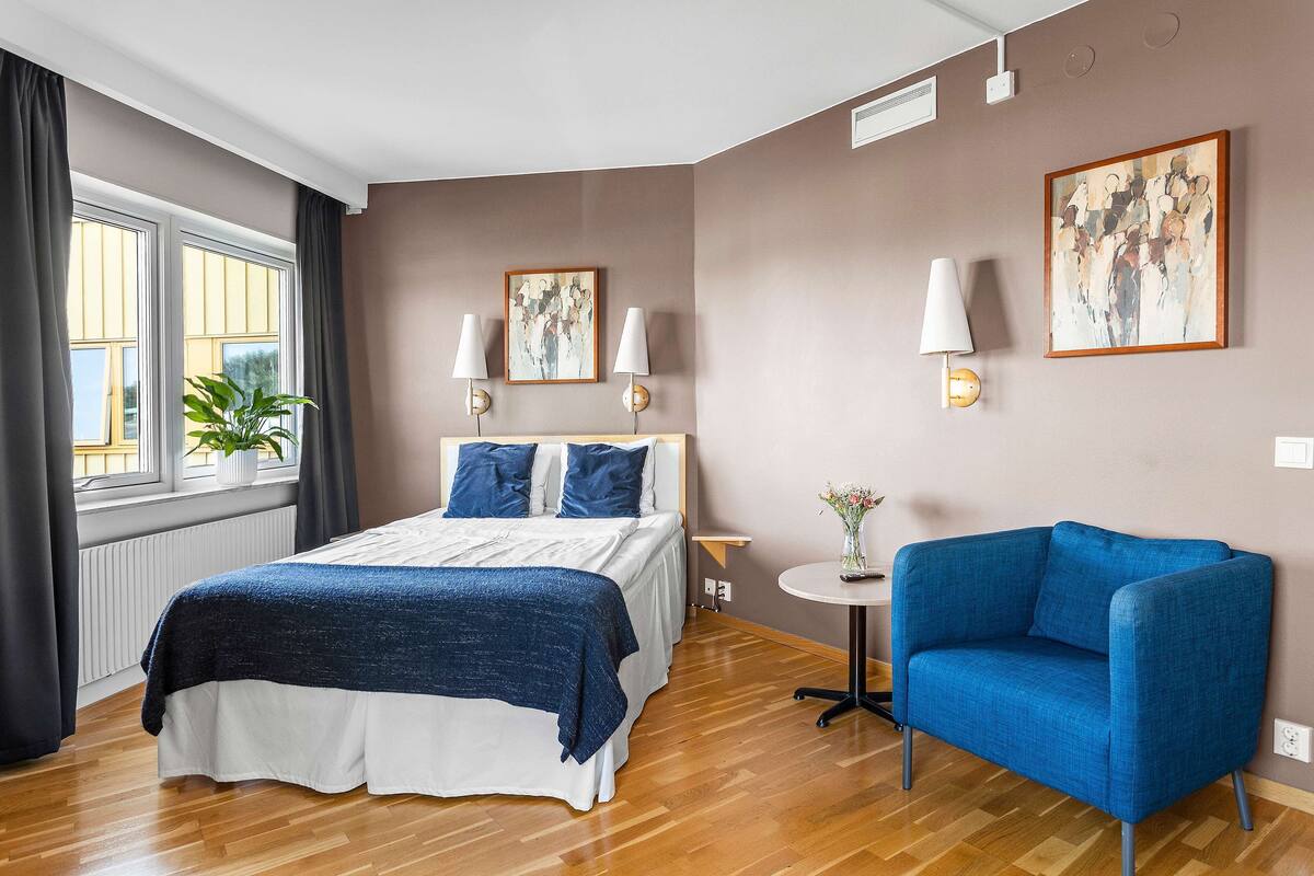 Une chambre à coucher moderne avec un lit, un fauteuil bleu, une petite table et des œuvres d’art accrochées aux murs.