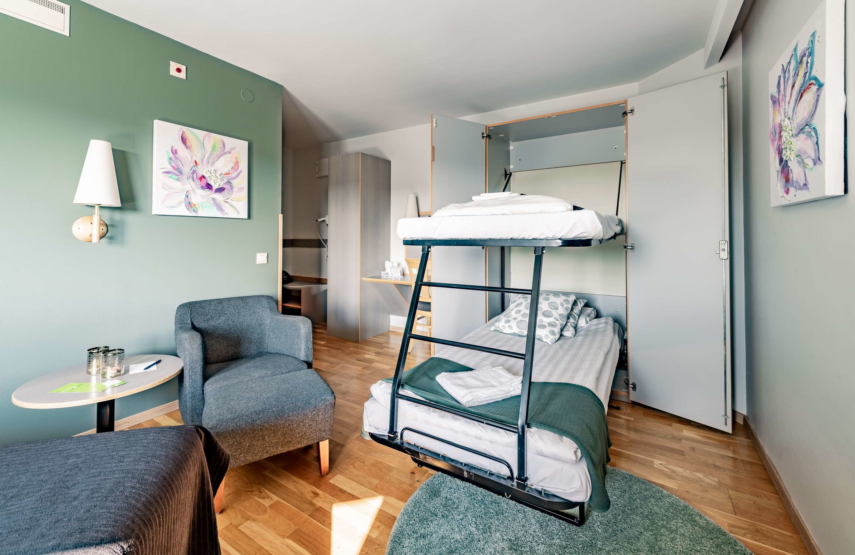 A compact hotel room with a bunk bed, a small desk with a lamp, a chair, and a round table.