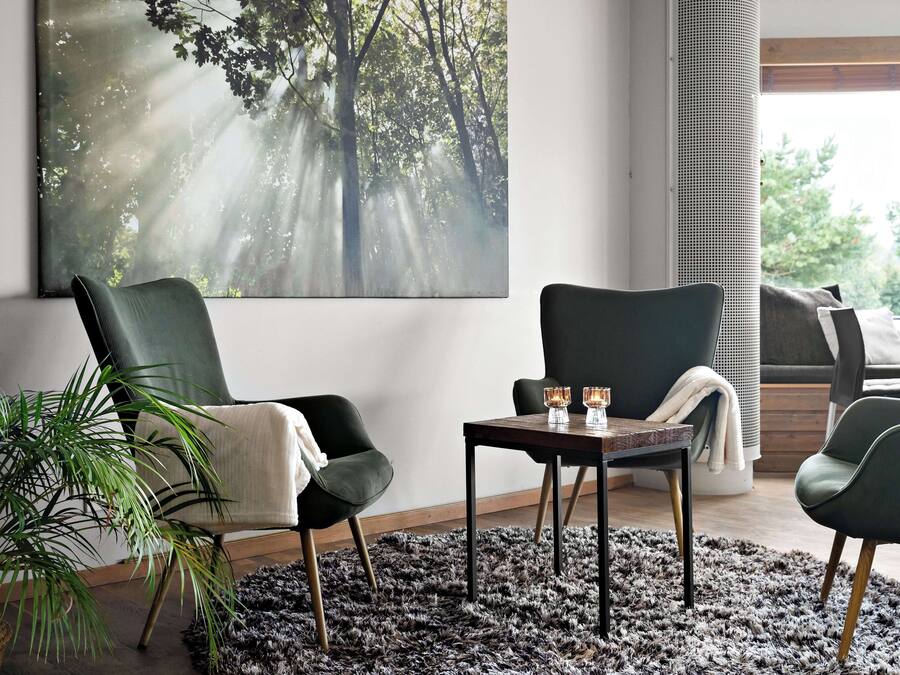 Una sala moderna con un cuadro de gran tamaño con temática forestal, dos sillones verde oscuro, una pequeña mesa de madera con dos velas y una planta en maceta.