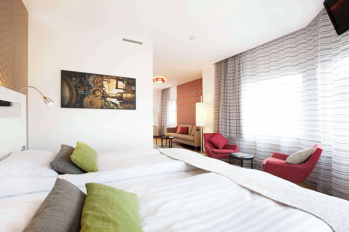 A hotel room with two beds, a red armchair, a sofa, and a painting on the wall.