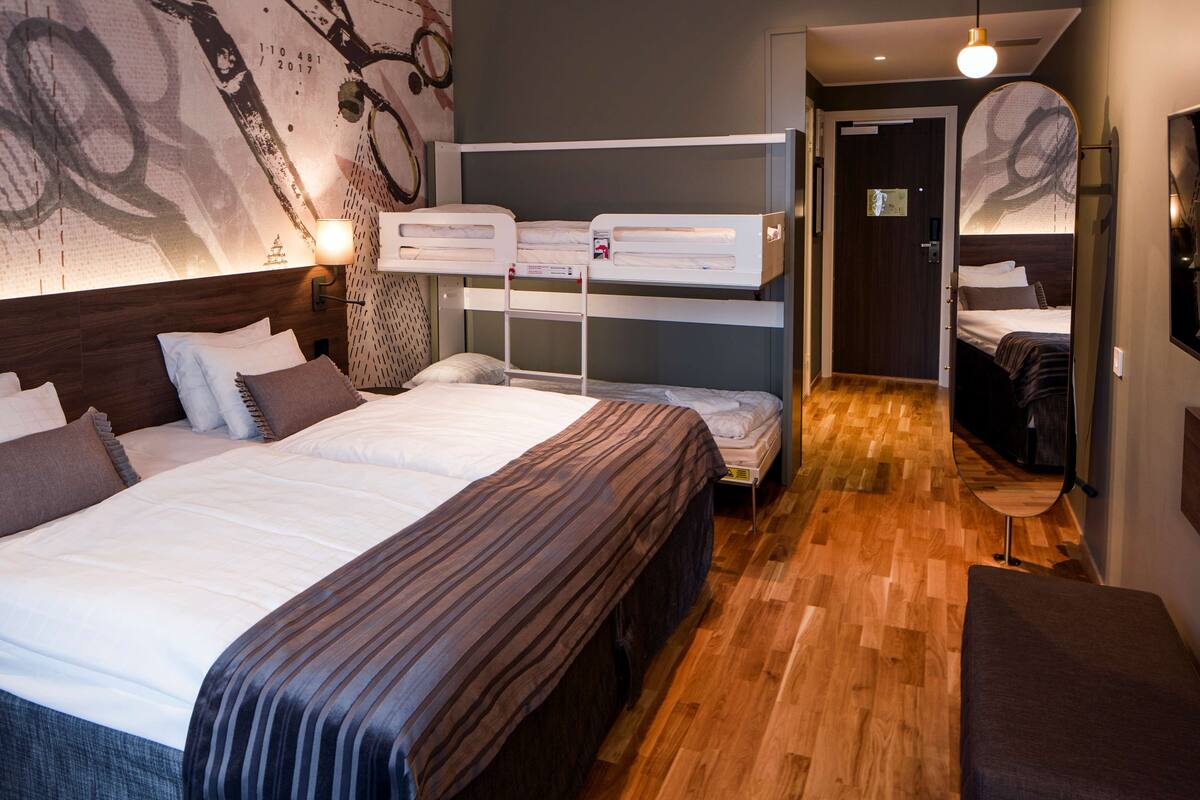 A modern hotel room with a large bed, a smaller bed on a platform, wooden flooring, and a mirror on the wall.