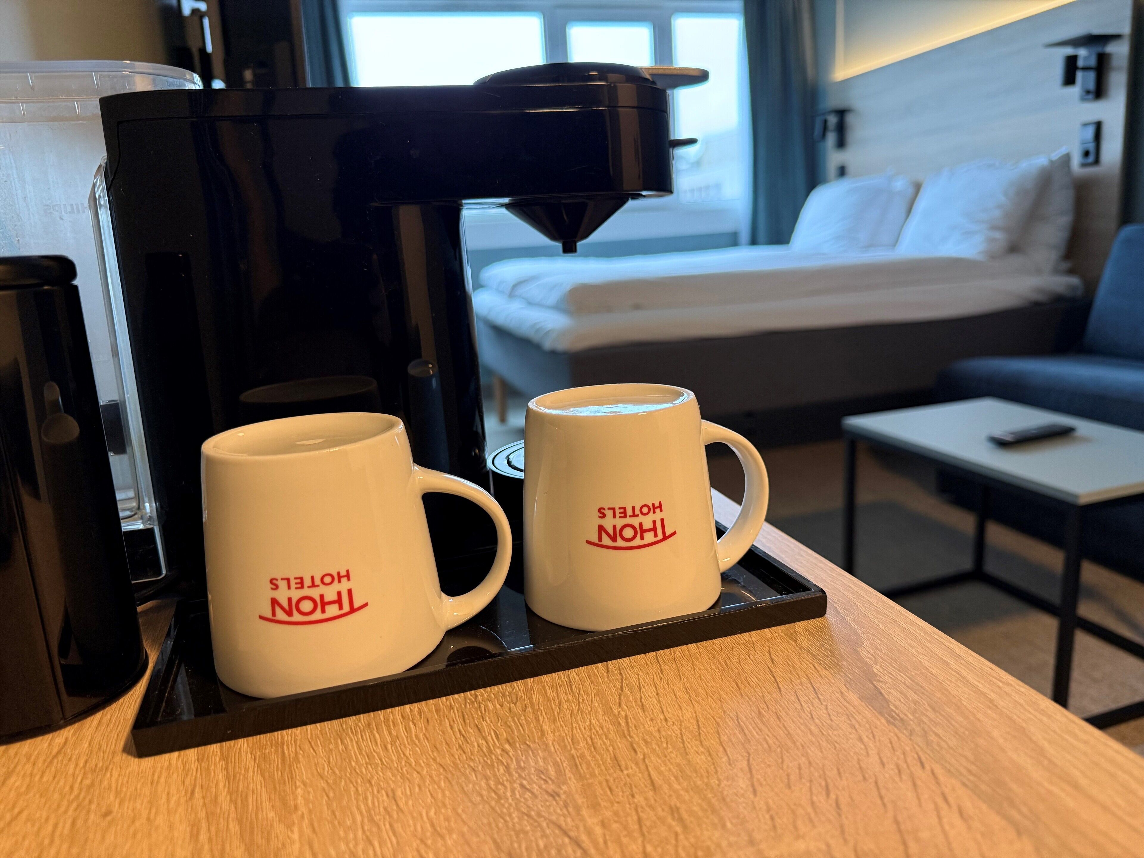 A hotel room with a coffee machine, two white mugs with red text, a bed, a sofa, and a small table.