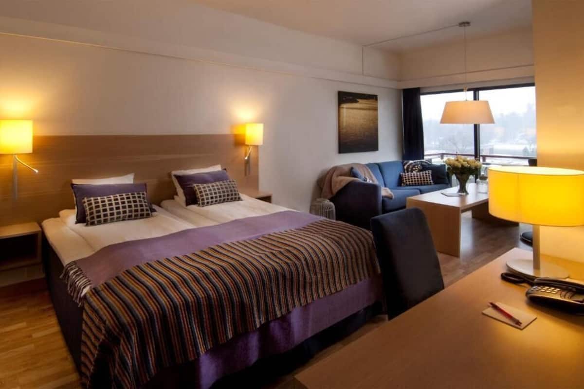 Habitación de hotel con cama, mesitas de noche, escritorio, una silla y vista a la ciudad por la ventana.