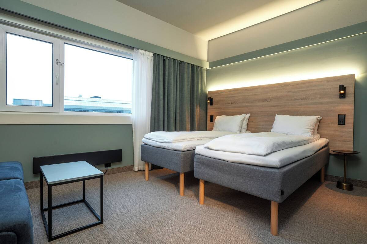 Quarto de hotel moderno com duas camas, uma mesinha, um sofá e vista para a cidade.