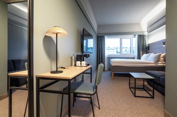 Habitación de hotel con cama, escritorio con lámpara, silla, mesita y ventana con cortinas.