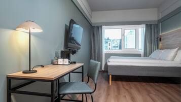 Habitación de hotel con cama, escritorio con lámpara, silla, televisor y ventana con cortinas.