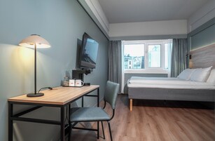 Habitación de hotel con cama, escritorio con lámpara, silla, televisor y ventana con cortinas.