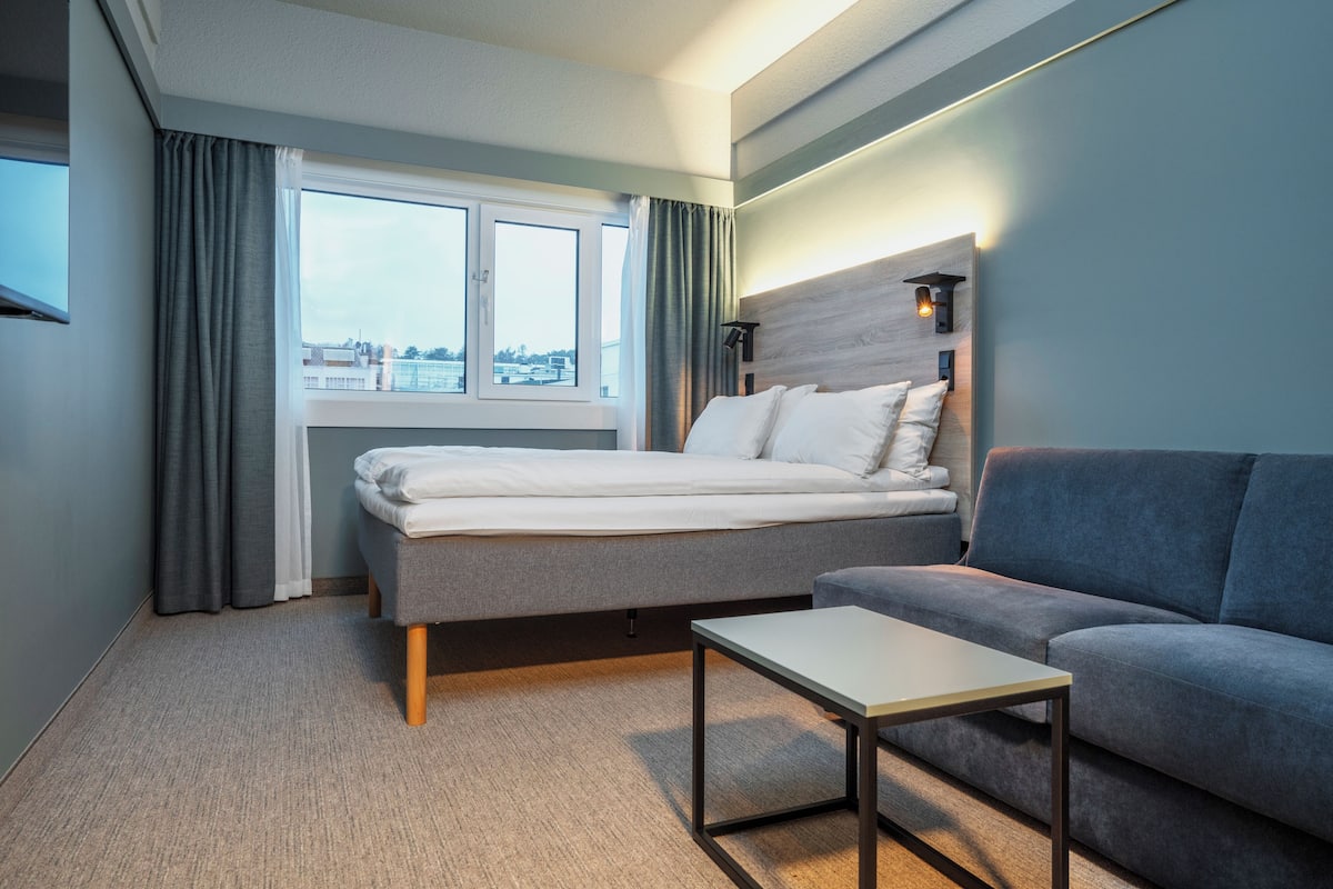 Quarto de hotel moderno com cama, sofá, mesinha e janela com cortinas.