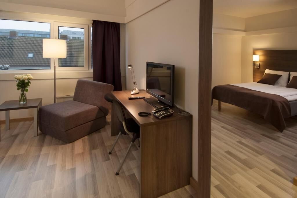 Habitación de hotel con escritorio, silla, televisión, cama y ventana con cortinas.